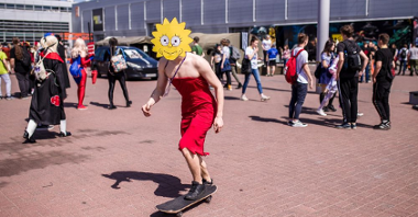 Uczestnik przebrany za postać Lisy Simpson z serialu animowanego "Simpsonowie" jedzie na deskorolce. Za nim uczestnicy Pyrkonu na dziedzińcu MTP.