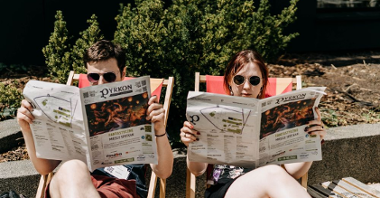 Chłopak i dziewczyna, oboje w przeciwsłonecznych okularach, siedzą na leżakach czytając gazetę festiwalową.