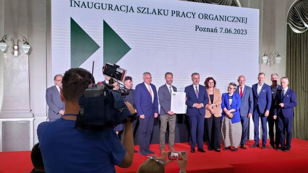 .Kilkanaście osób: mężczyzn i kobiet na scenie, za nimi tablica z napisem Inaugracja Szlaku Pracy Organicznej i data, widok częściowo przesłania operator kamery filmujący ludzi na scenie.
