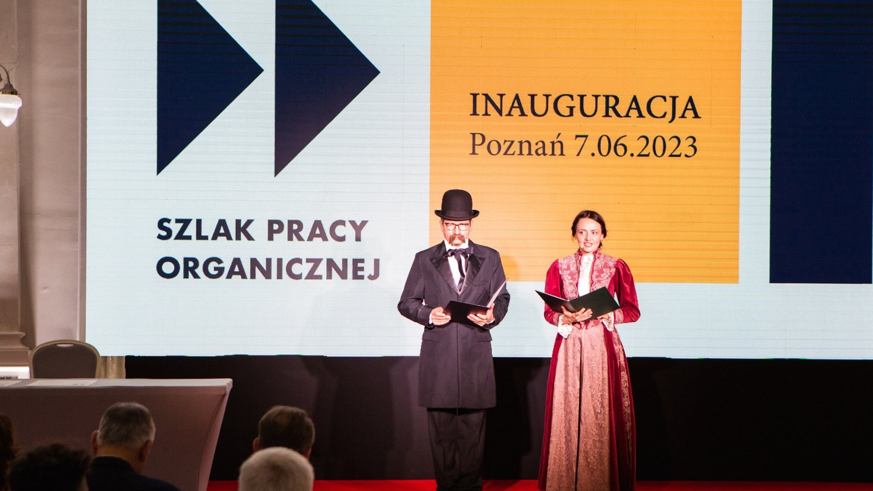 Mężczyzna i kobieta w strojach stylizowanych na XIX wiek odczytują nazwiska osób zaangażowanych w projekt