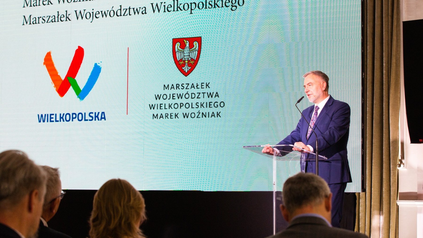 Na scenie przy mównicy mężczyzna w granatwym garniturze, za nim plansza z jego nazwiskiem i zajmowanym stanowiskiem