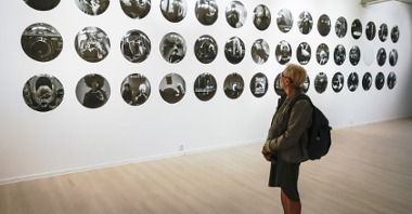 Na białej ścianie wisi dużo małych, okrągłych, czarno-białych zdjeć przedstawiających Izabellę Gustowską. Ogląda je uczestniczka wernisażu.