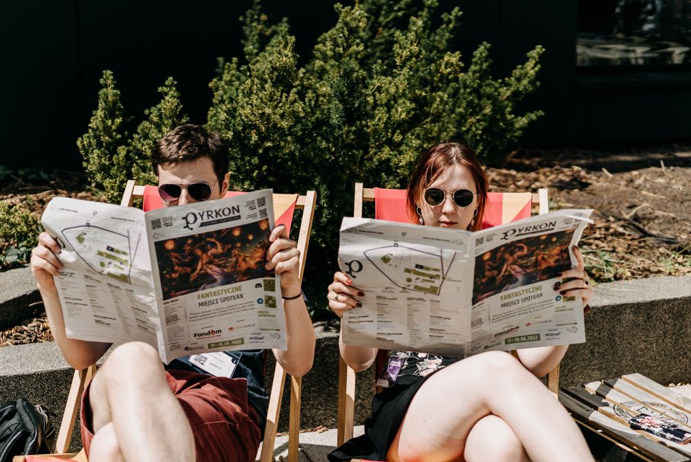 Chłopak i dziewczyna, oboje w przeciwsłonecznych okularach, siedzą na leżakach czytając gazetę festiwalową. - grafika artykułu