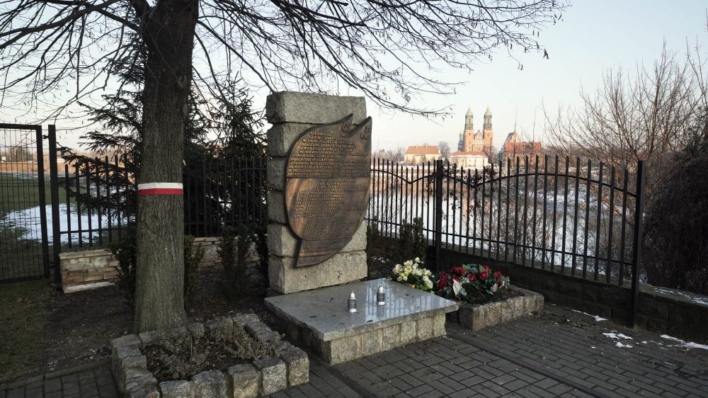 Obok pomnika stoją dwa znicze i wazon pełen kwiatów, jest osłonięty drzewem. W oddali, za pomnikiem, widać katedrę. Zdjęcie robione zimą, co widać po leżącym gdzieniegdzie śniegu.