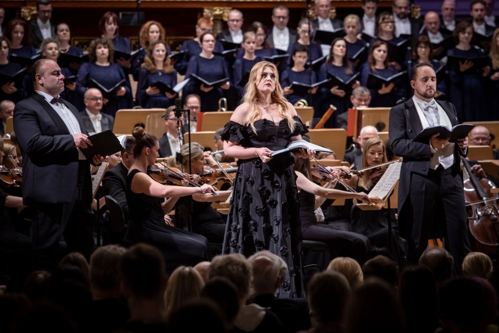 W centrum solistka ubrana w elegancką czarną suknię z tłoczonymi kwiatami, w rozpuszczonych blond włosach, po obu jej stronach dwóch męskich solistów, za nią orkiestra i chór. - grafika artykułu