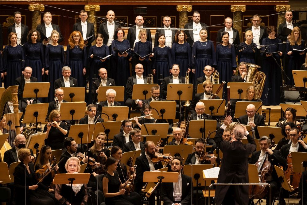 U dołu zdjęcia dyrygent, nad nim widać orkiestrę, sekcję smyczkową oraz instrumentów dętych, nad nimi, w dwóch rzędach, chór. - grafika artykułu