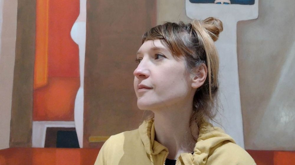 Młoda kobieta w żółtej bluzie i jasnymi włosami spiętymi w luźny kok. Patrzy w przestrzeń. Za nią malarskie tło.