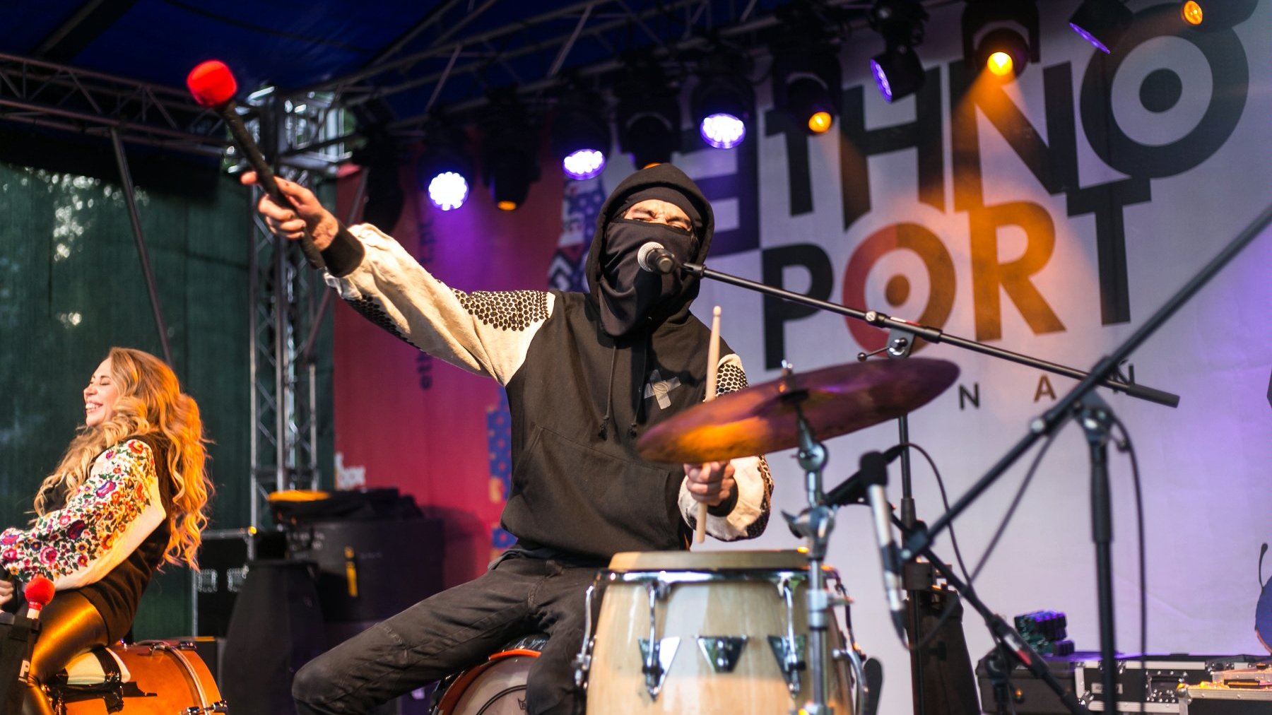 Artysta występuje na scenie. Siedzi, gra na bębnach i śpiewa do mikrofonu. Ma ubraną kominiarkę i bluzę. Jedną rękę trzyma w górze. Za nim siedzi artystka.
