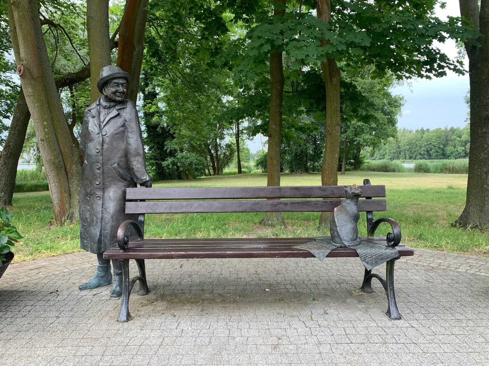 Uliczny pomnik w formie ławki, przy której po jednej stronie stoi podobizna Wisławy Szymborskiej, a po drugiej - kot. - grafika artykułu