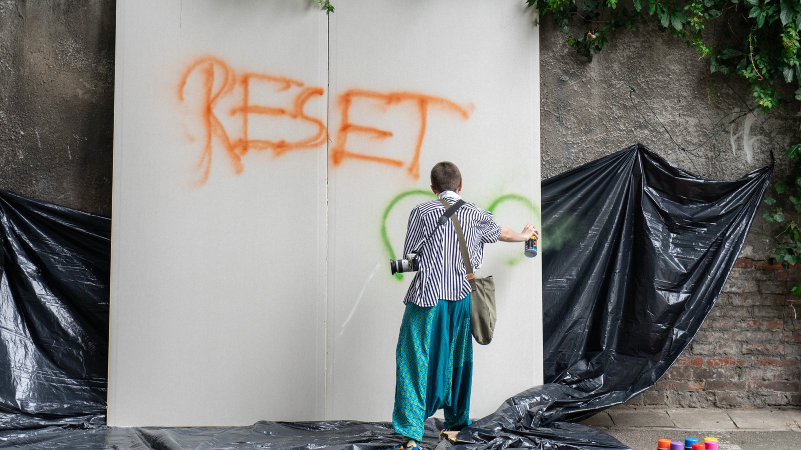 Dziewczyna stoi tyłem do obiektywu i sprayem rysuje napis "RESET" na białym płótnie.