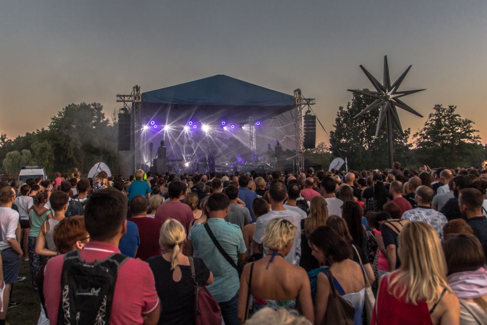 Publiczność patrzy w kierunku rozświetlonej, plenerowej sceny, na której odbywa się koncert. - grafika artykułu