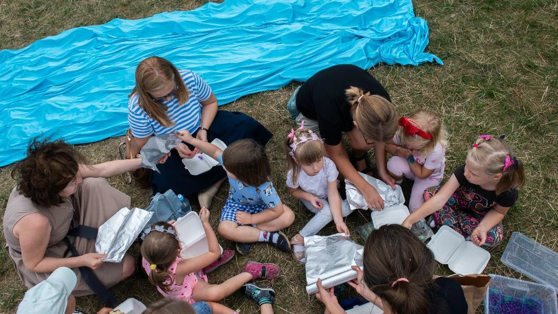 Plener. Widok z lotu ptaka. Grupa dzieci i dorosłych siedzi na trawie i pakuje do opakowań jagody. Za nimi niebieski materiał, symbolizujący rzekę.