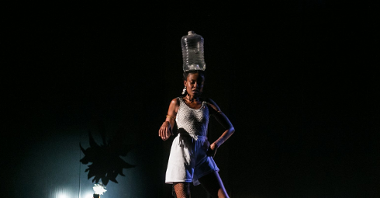 Kobieta w białym stroju i z plastikową butlą na głowie tańczy wśród rozsypanego cukru. Stoi na zgiętych nogach i wystawia jedną rękę w stronę obiektywu.