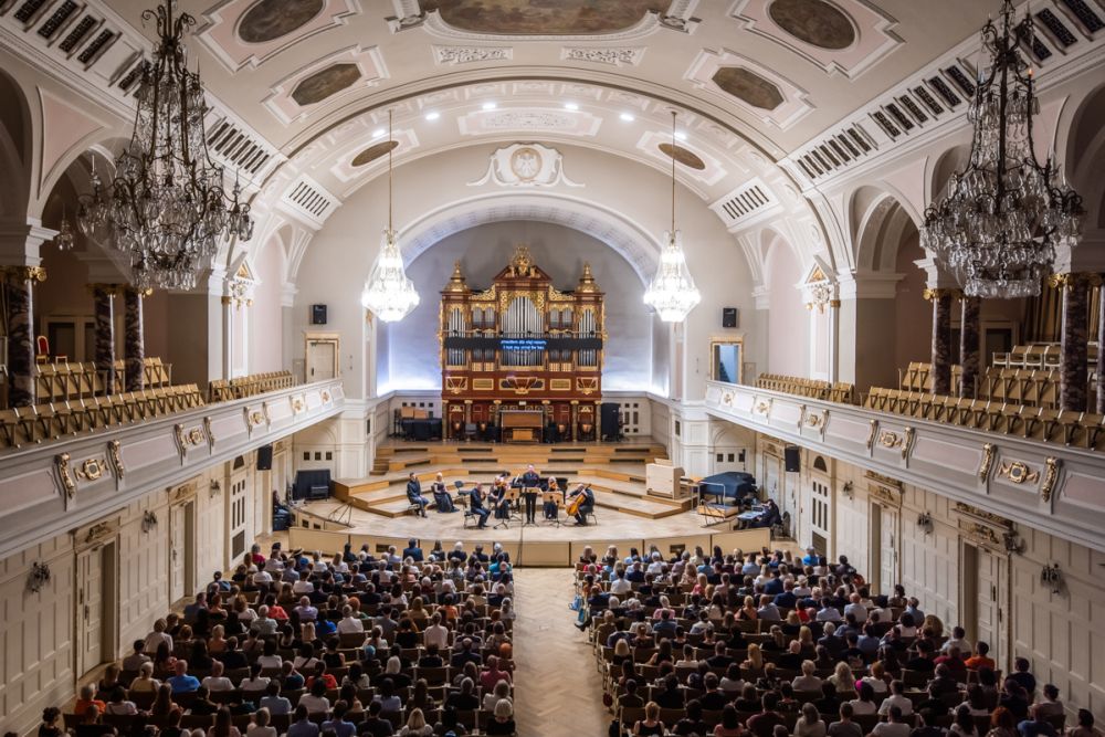 Widok na Aulę UAM z góry. Na scenie widać występujących muzyków, na fotelach zasiada publiczność. Na wszystkim górują zabytkowe, złocone organy. - grafika artykułu