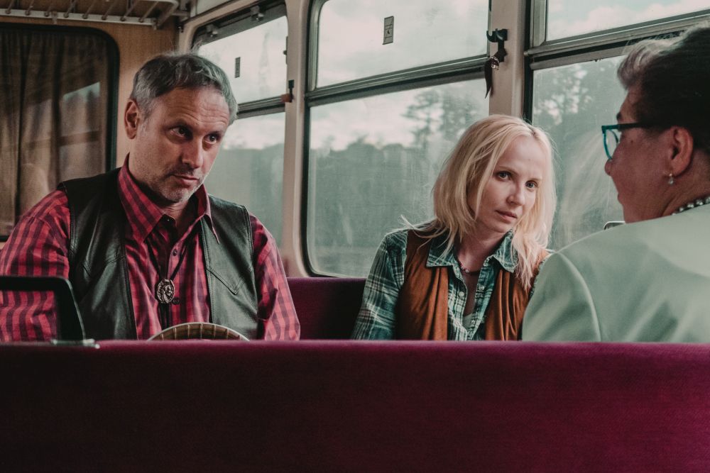Jasnowłosa kobieta i mężczyzna w czerwonej, kraciastej koszuli siedzą w pociągu, w wagonie bezprzedziałowym. Patrzą uważnie na siedzącą na przeciwko starszą kobietę w okularach. - grafika artykułu