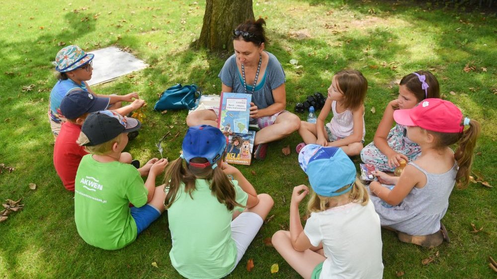 Dzieci siedzą na trawie wokół dorosłej opiekunki, która pokazuje im książkę o podróżowaniu po świecie. Jest pogodny ciepły dzień.