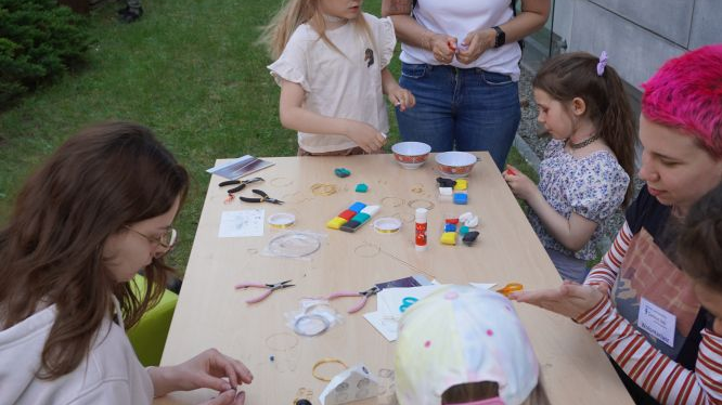 Dzieci oraz ich opiekunowie stoją wokół stołu zastawionego farbami, nożyczkami, cążkami, plasteliną i wykonują zadania manualne.