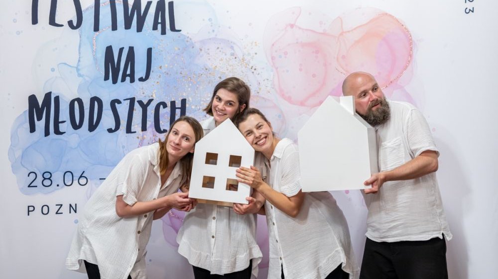 Trzy dziewczyny i jeden mężczyzna w białych koszulkach, pozują, trzymając białe, drewniane domki, udając, że nasłuchują. Za nimi plakat Festiwalu Najmłodszych.