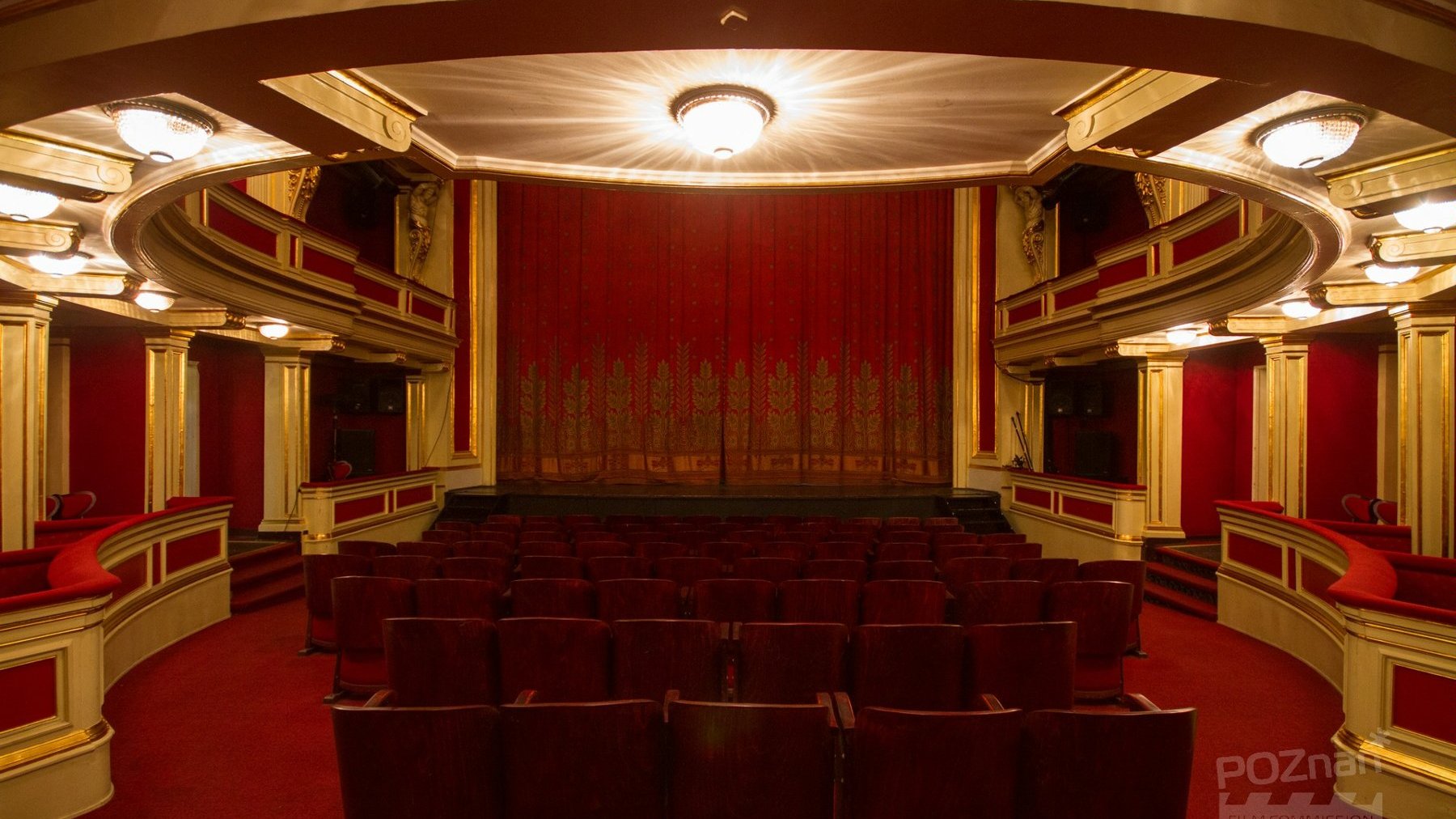 Sala teatralna, widok na scenę zasłonietą kurtyną. Czerwone krzesła i ściany ze złotymi elementami. W górze żyrandol.
