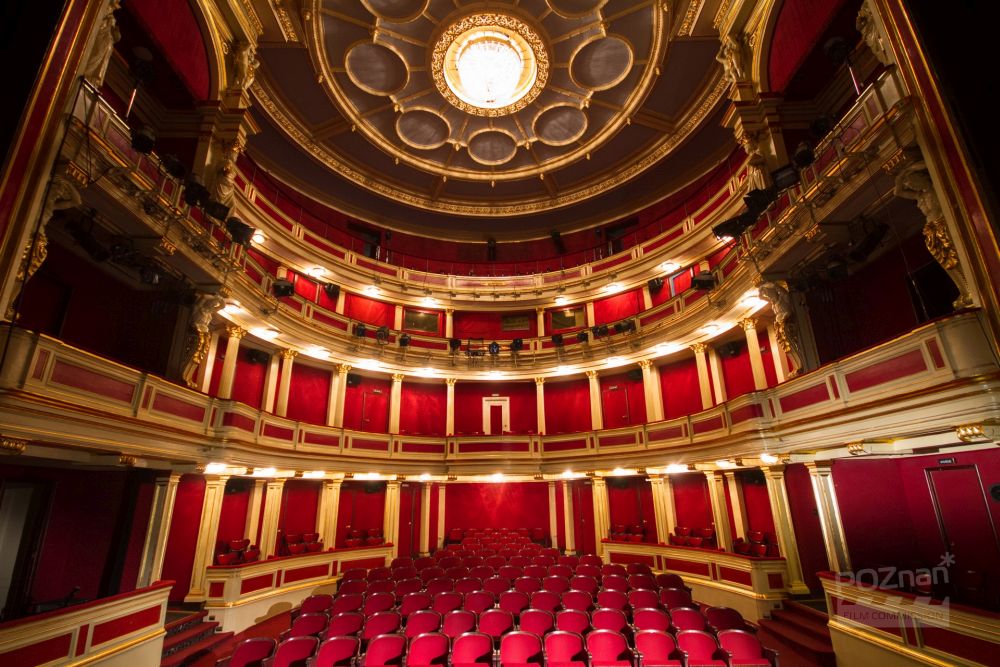 Sala teatralna, widok na publiczność. Czerwone krzesła i ściany ze złotymi elementami. W górze żyrandol. - grafika artykułu