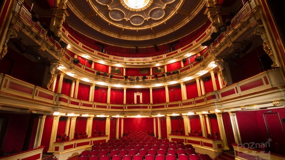 Sala teatralna, widok na publiczność. Czerwone krzesła i ściany ze złotymi elementami. W górze żyrandol.