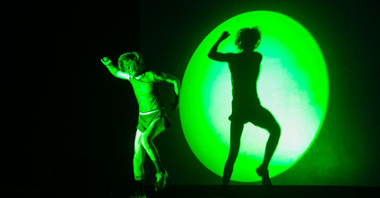 Podświetlony na zielono performer tańczy. Jego cień na ścianie odzwierciedla jego ruchy.