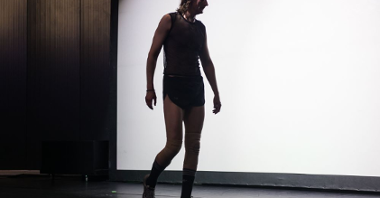 Tancerz ubrany w koszulkę, krótkie spodenki, skarpetki i sportowe buty, idzie przez scenę, ma opuszczone ręce i ramiona.