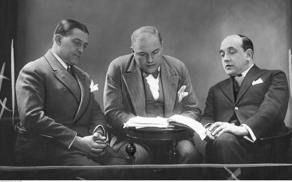 Czarno-biała fotografia. Trzech ubranych elegancko mężczyzn siedzi przy okrągłym stoliku, wpatrują się w leżący na nim plik kartek. - grafika artykułu