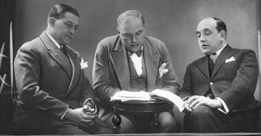 Czarno-biała fotografia. Trzech ubranych elegancko mężczyzn siedzi przy okrągłym stoliku, wpatrują się w leżący na nim plik kartek.