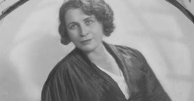 Czarno-biała fotografia. Kobieta w długiej sukni pozuje do zdjęcia siedząc. Na lewej ręce nosi bransoletkę.