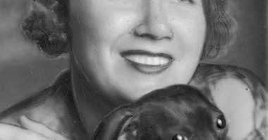 Zdjęcie portretowe uśmiechniętej kobiety. Obiema rękami przytula do siebie czarnego jamnika, pies spogląda prosto w obiektyw.
