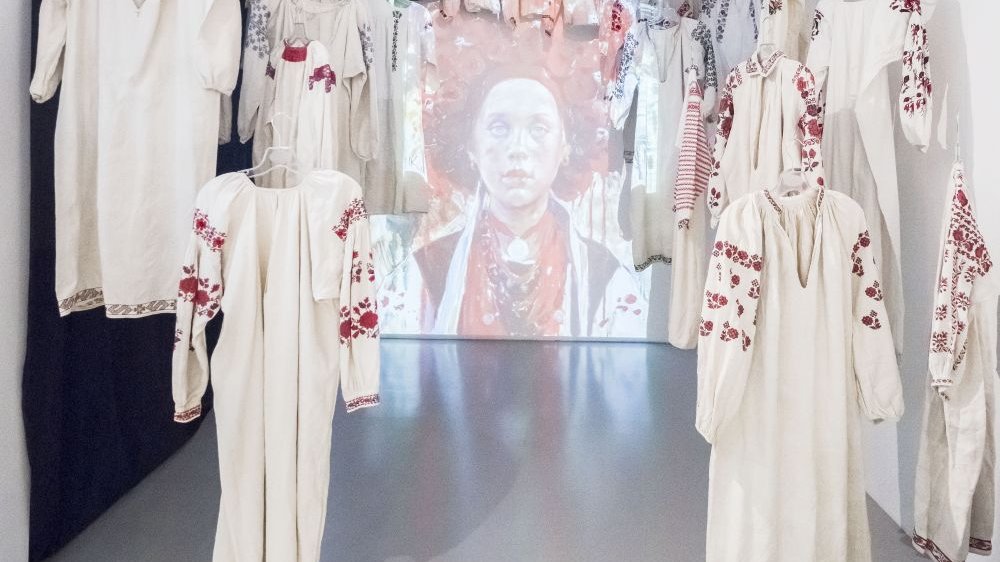 Na środku ekran z reprodukcją portretu kobiety w ludowym stroju, dookoła niego zwisające z sufitu stroje ludowe. Kadr utrzymany w bieli z elementami czerwieni.