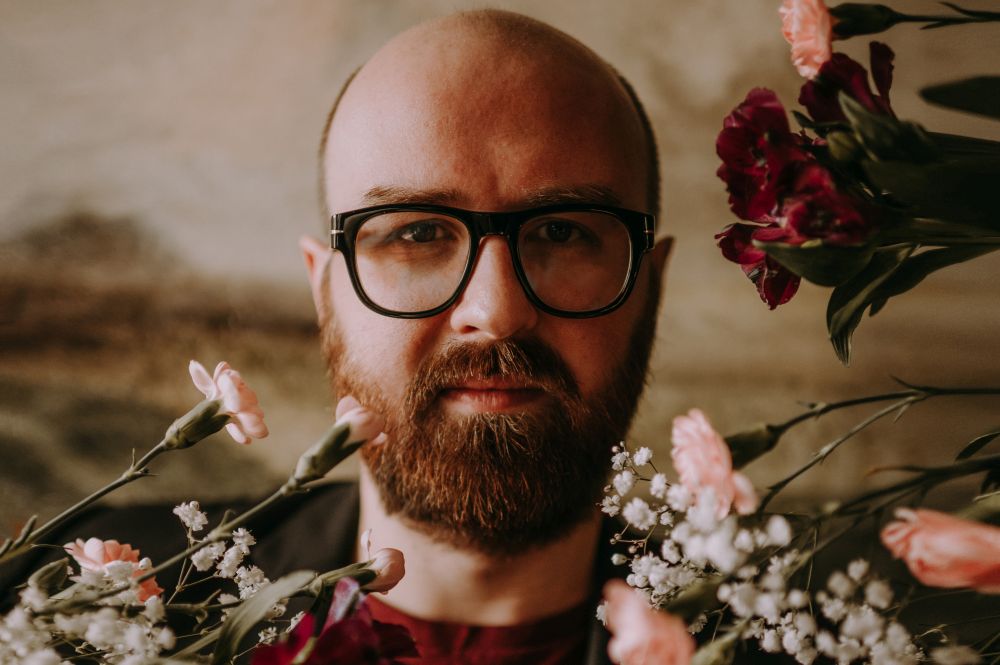 Portret mężczyzny w okularach i z brodą w otoczeniu kwiatów. - grafika artykułu