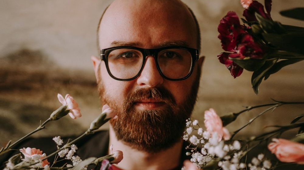 Portret mężczyzny w okularach i z brodą w otoczeniu kwiatów. - grafika artykułu