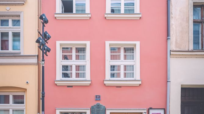 Trzypiętrowa wąska kamienica w kolorze różowym z białymi okiennicami w sąsiedztwie innych budynków.