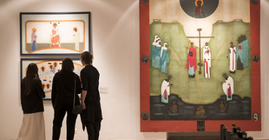 Trzy osoby ubrane w ciemne kolory oglądają obrazy na wystawie. Stoją przy mniejszym, białym obrazie w czarnych ramach.
