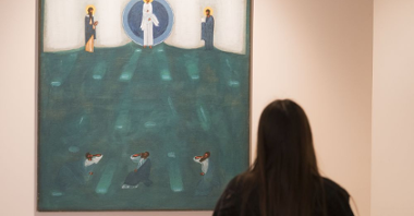 Zwiedzająca wystawę dziewczyna przygląda się jednej z ikon - dużej, na której namalowane zostało wyobrażenie biblijnej sceny. Dominujące kolory to niebieski i zielony.