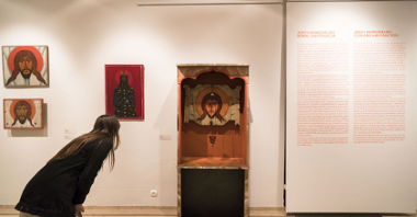 Zwiedzająca wystawę pochyla się obok jednej z ikon, umieszczonej w głębi drewnianego regału. Obok wiszą inne, małe ikony oraz opis wystawy.