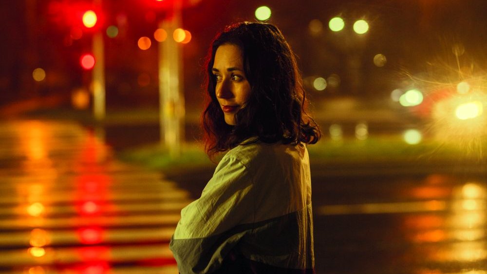 Dziewczyna w ciemnych włosach spogląda w lewo. Stoi przed przejściem dla pieszych, w świetle ulicznych świateł. Jest ciemno, asfalt jest mokry od deszczu. - grafika artykułu