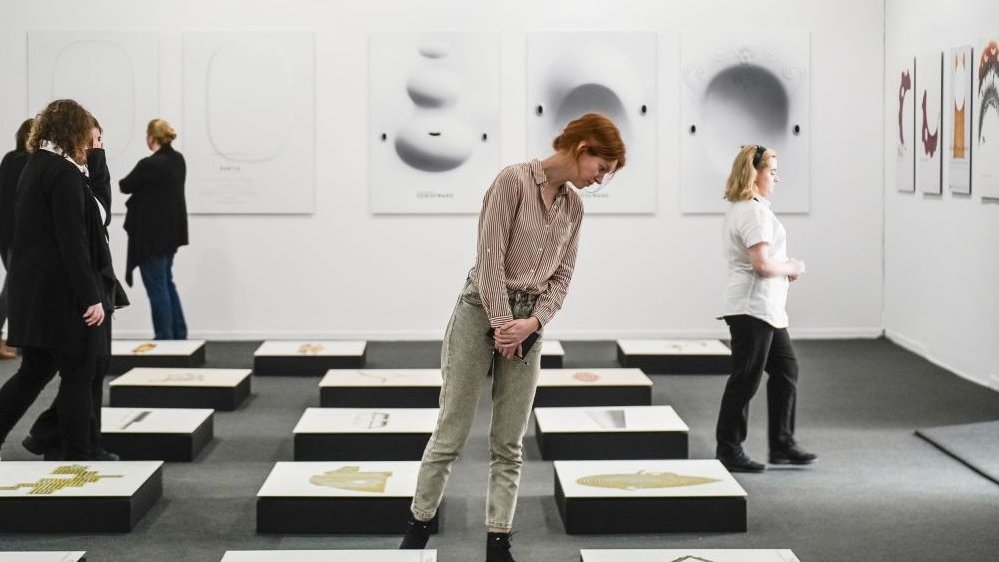 Ludzie zwiedzający wystawę oglądają obiekty leżące na podłodze i grafiki wiszące na ścianach. Sala utrzymania w kolorze bieli.