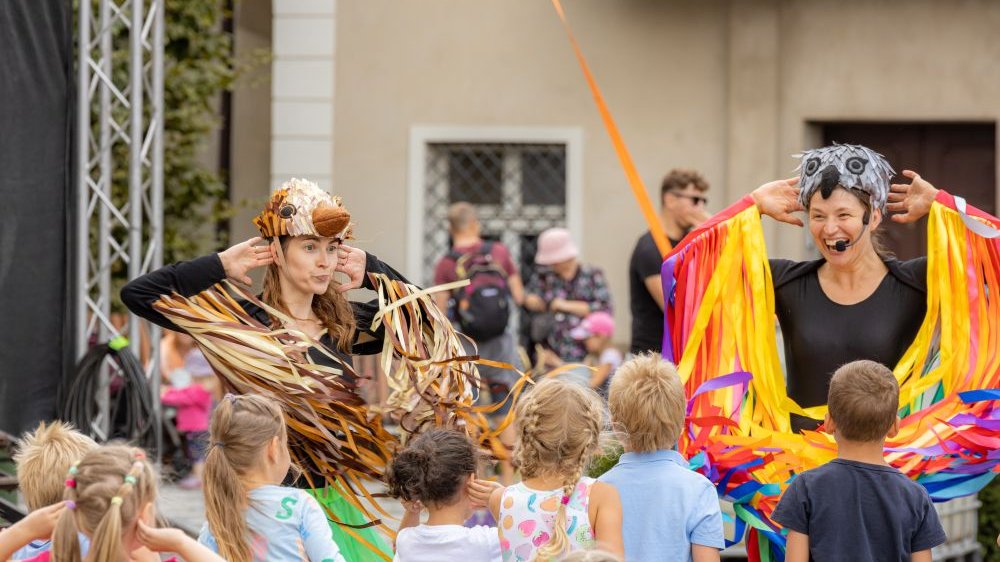 Dwie aktorki dziecięcego teatru występują przed małą publicznością. Obie są przebrane za ptaki, ręce mają uniesione za głowę.