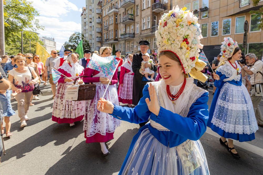 Barwny pochód ludzi ubranych w tradycyjne stroje poznańskie. Dziewczyna na pierwszym planie uśmiecha się szeroko, macha do przechodniów. - grafika artykułu