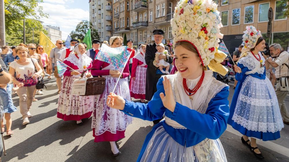 Barwny pochód ludzi ubranych w tradycyjne stroje poznańskie. Dziewczyna na pierwszym planie uśmiecha się szeroko, macha do przechodniów.
