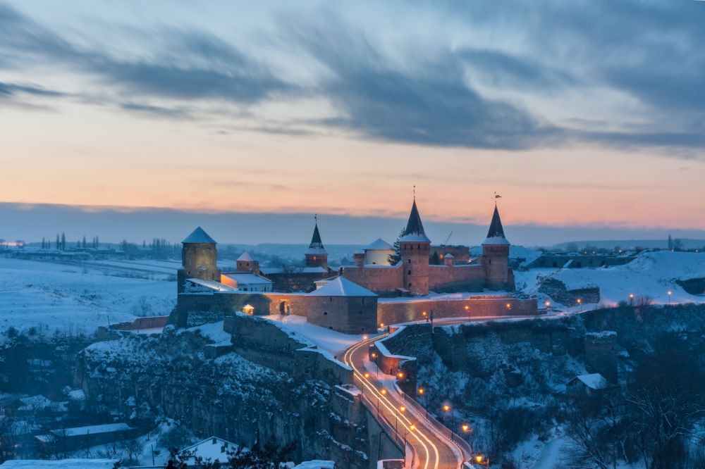 Zimowy wieczorowy krajobraz. W centrum zdjęcia ogromny średniowieczny zamek, bardzo dobrze zachowany, górujący nad miasteczkiem, subtelnie oświetlony światełkami latarni. - grafika artykułu