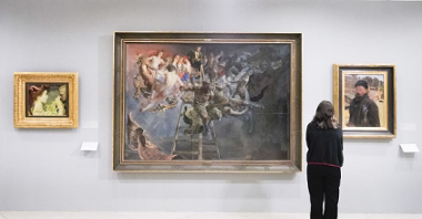 Obrazy Jacka Malczewskiego na ścianie Muzeum, ogląda je kobieta w czerni. Obraz na środku zdecydowanie góruje rozmiarami nad pozostałymi obrazami po swojej prawej i lewej stronie.