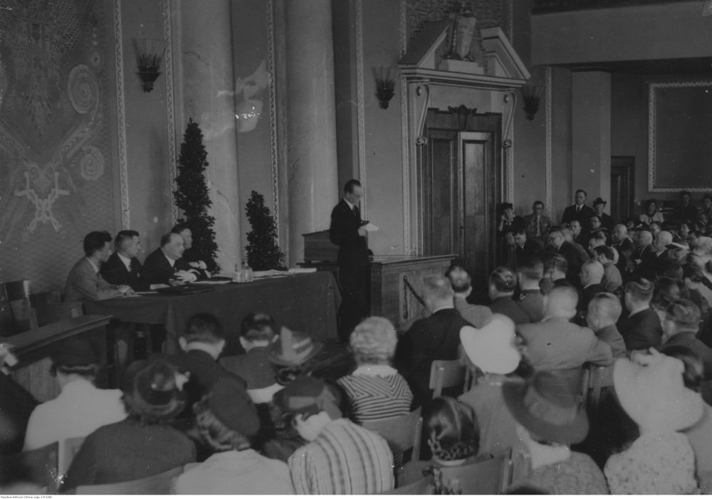 Czarno-biała fotografia mężczyzny w garniturze przemawiającego do sali pełnej ludzi. Z tyłu, za jego plecami, czterech innych mężczyzn siedzi za stołem. - grafika artykułu