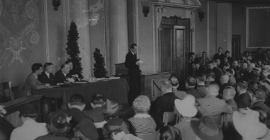 Czarno-biała fotografia mężczyzny w garniturze przemawiającego do sali pełnej ludzi. Z tyłu, za jego plecami, czterech innych mężczyzn siedzi za stołem.