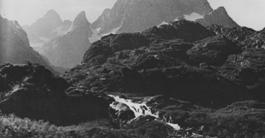 Czarno-biała fotografia norweskiego fiordu z wodospadem. W tle wysokie góry, w powietrzu unosi się mgła.