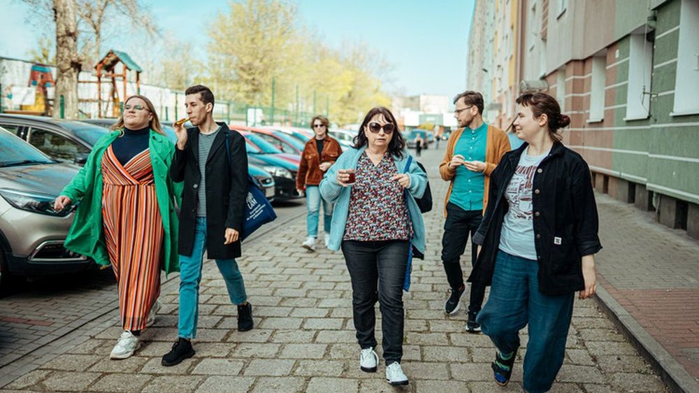 Grupa kolorowo ubranych ludzi spaceruje ulicą. Po lewej parking samochodowy, za nim dziecięcy plac zabaw.