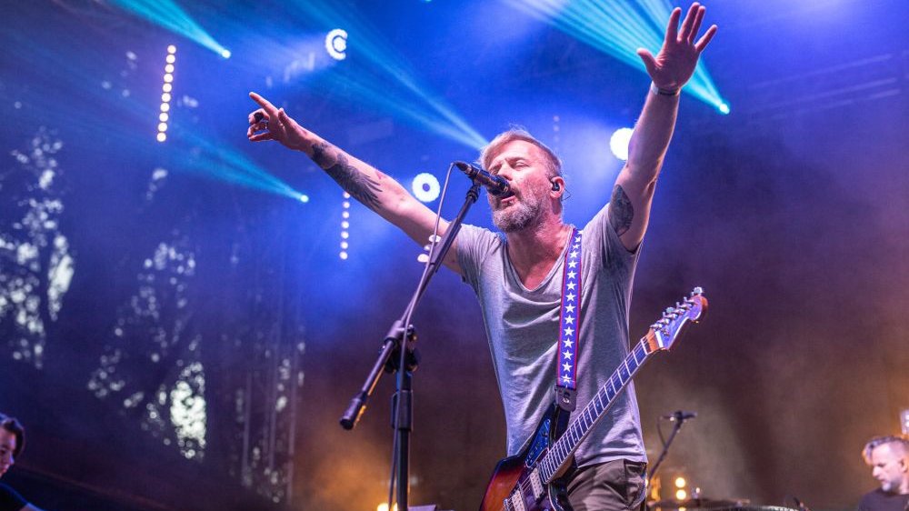 Organek śpiewa na scenie z gitarą przewieszoną przez ramie, wyciąga ręce do fanów pod sceną. Za nim reflektory świecące na niebiesko.